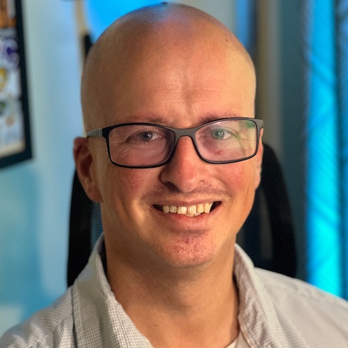 Smiling headshot of a bald bespectacled man wearing a collared button-up shirt. He's got a blue hue being displayed behind him on the wall